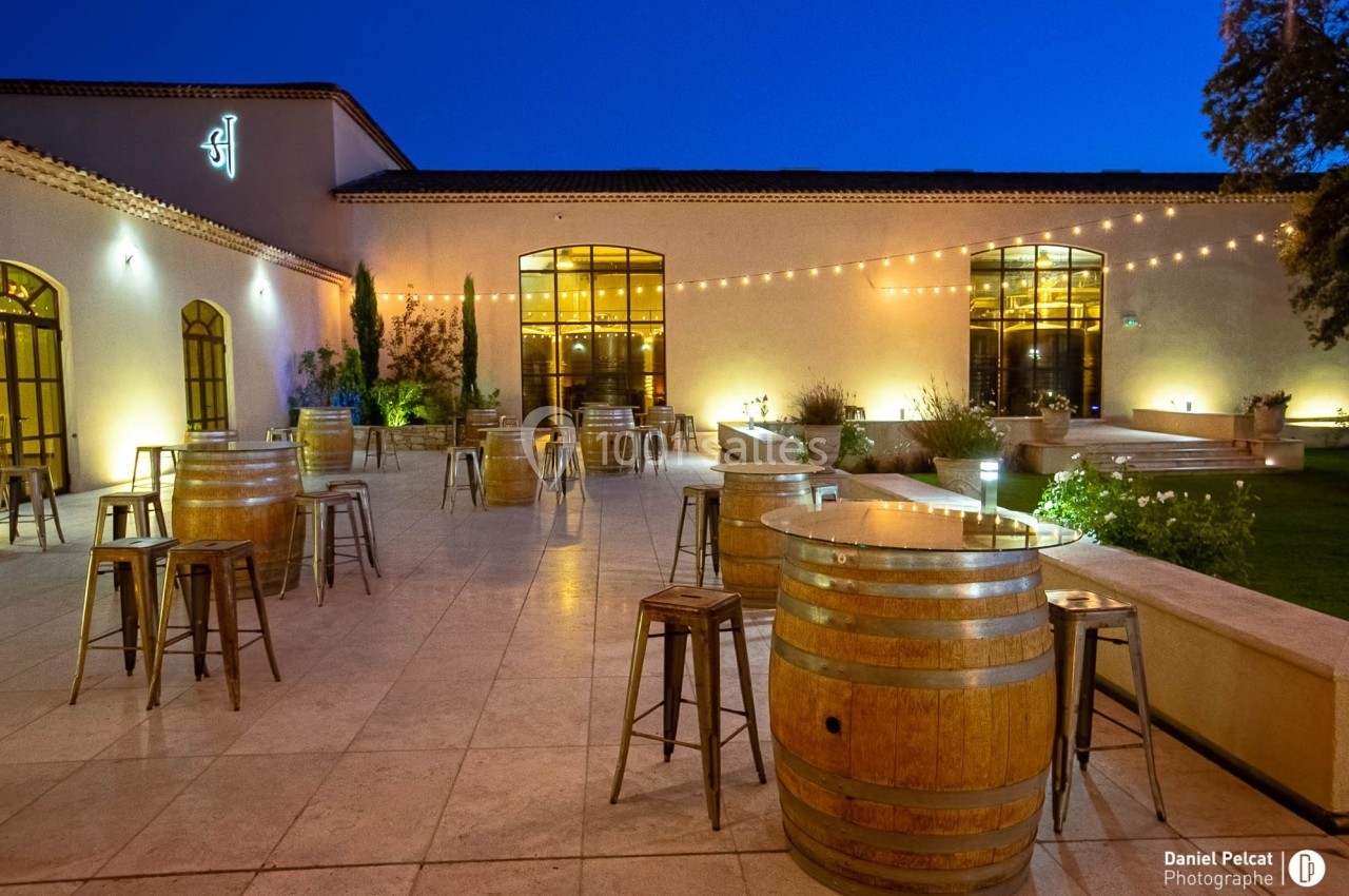 Terrasse éclairée en soirée avec tables en tonneaux, chaises métalliques et guirlandes lumineuses suspendues.