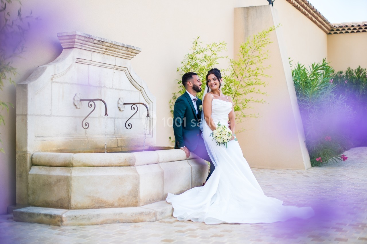 Un couple de mariés pose près d'une fontaine en pierre, entouré de végétation et de fleurs violettes floues au premier plan.