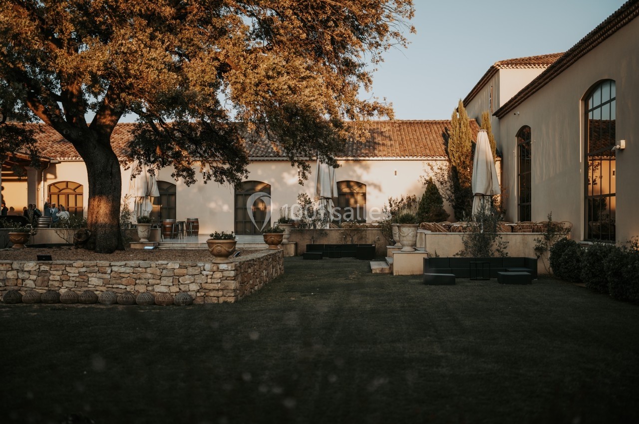 Cour extérieure d'une maison en pierre avec terrasse, parasols, arbres et pelouse au coucher du soleil.