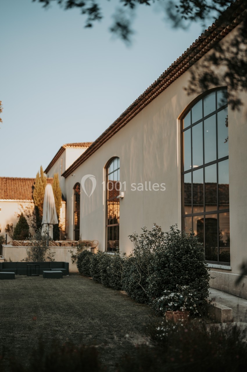Façade d'une maison avec grandes fenêtres, jardin entretenu et parasol sur une terrasse au coucher du soleil.
