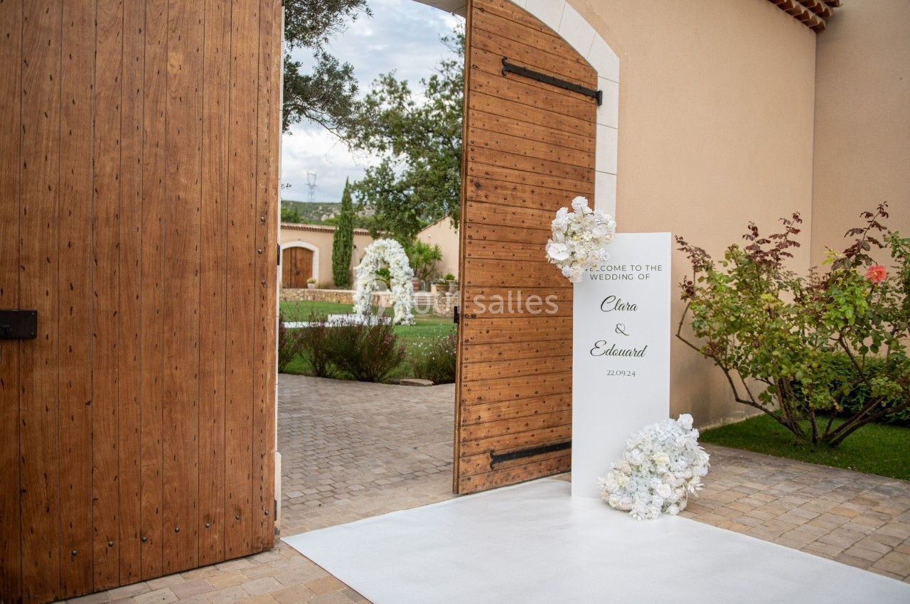 Entrée d'un lieu de mariage avec une porte en bois ouverte, une allée blanche et une décoration florale blanche.