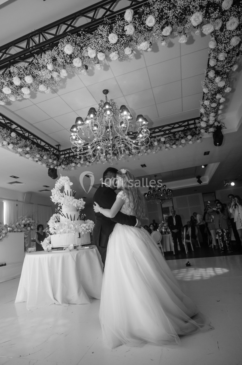 Un couple danse sous un lustre dans une salle décorée de fleurs, près d'un grand gâteau de mariage.