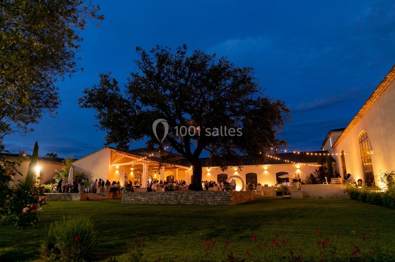 Cour extérieure éclairée par des guirlandes lumineuses, avec un grand arbre central et des invités rassemblés en soirée.