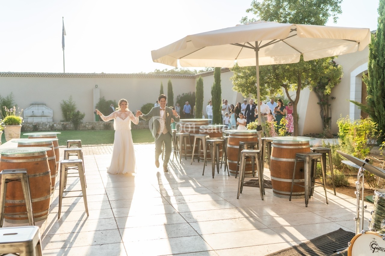 Un couple en tenue de mariage entre sur une terrasse ensoleillée décorée de tonneaux, entouré d'invités.