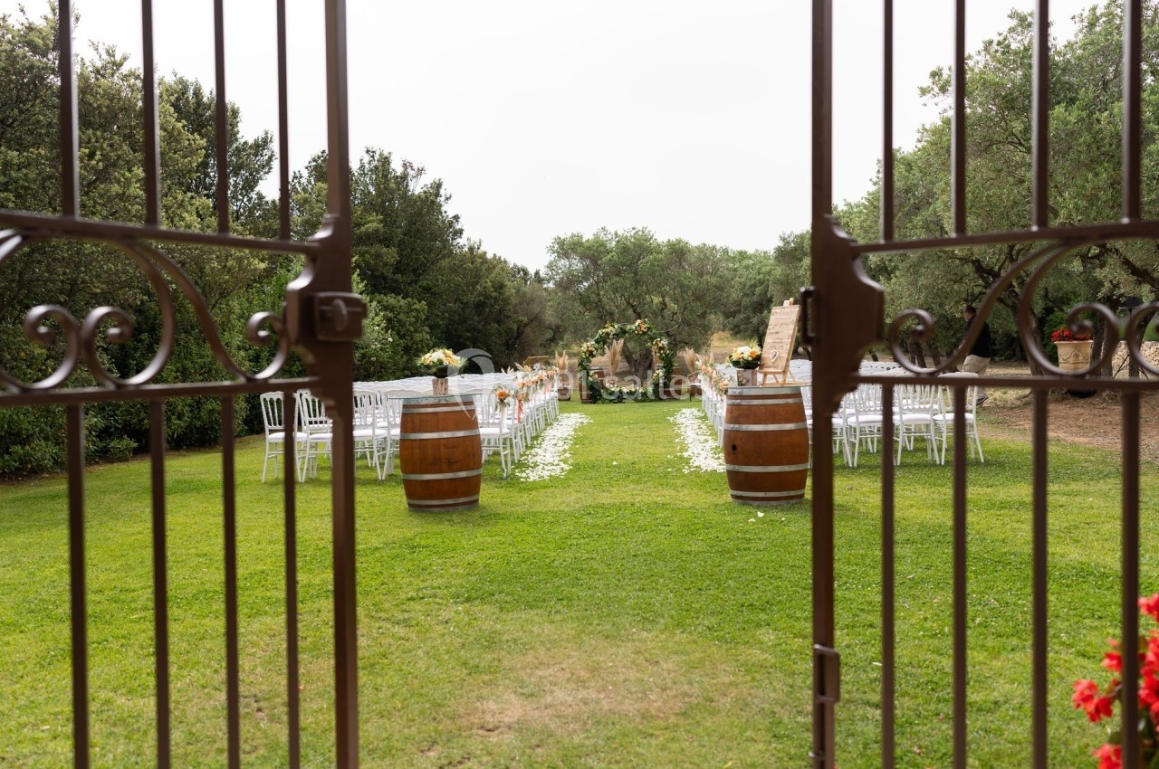 Allée centrale bordée de chaises blanches et de tonneaux décorés, menant à une arche fleurie dans un jardin verdoyant.