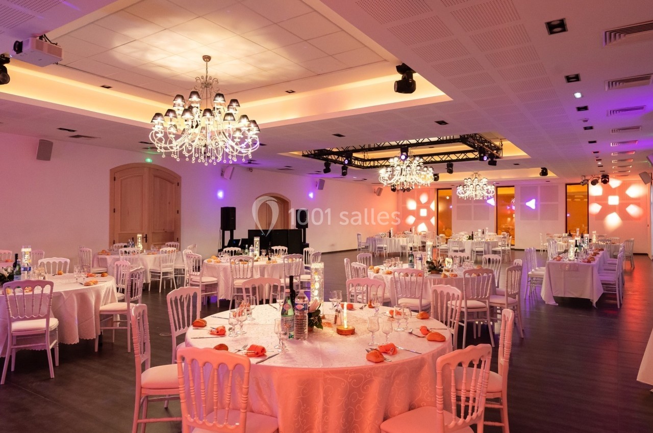 Salle de réception élégante avec tables rondes dressées, chandeliers suspendus et éclairage tamisé.
