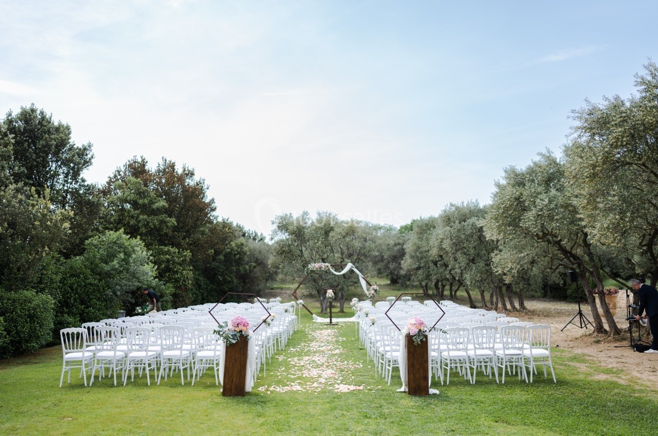 Chaises blanches alignées en extérieur, formant une allée centrale menant à une arche décorée pour une cérémonie.