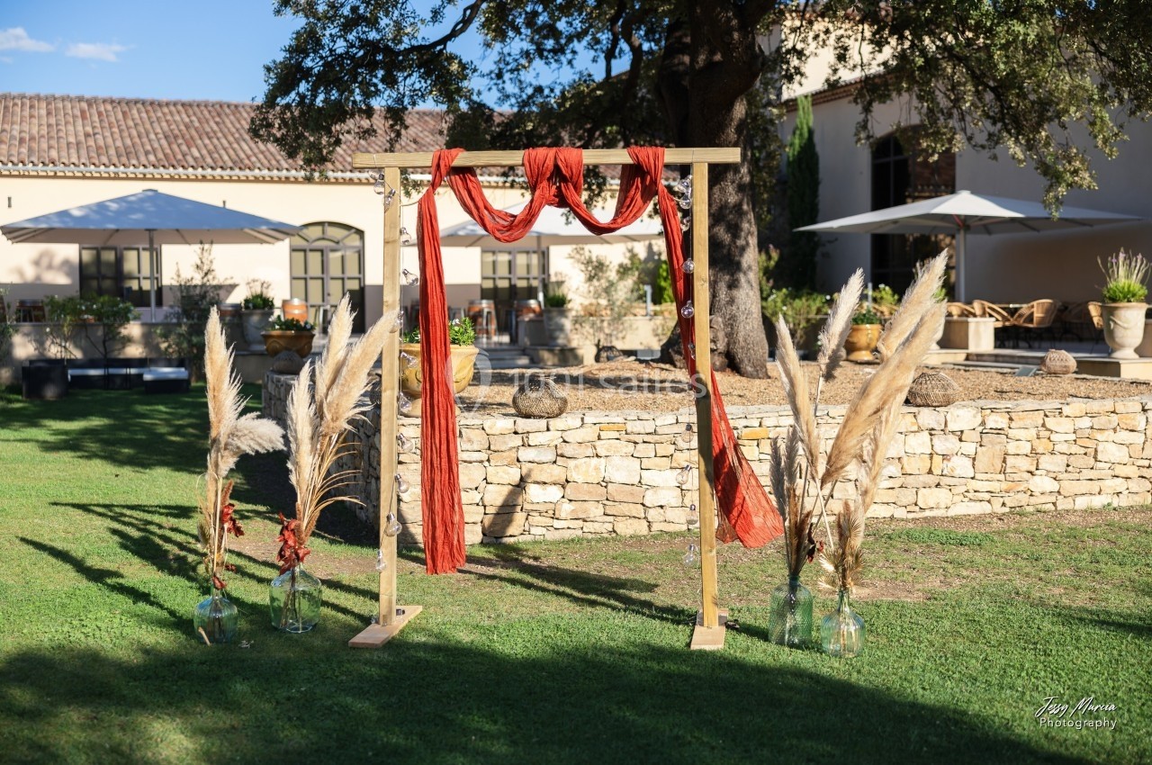 Arche en bois décorée de tissu rouge et de pampas, installée sur une pelouse devant un bâtiment en pierre.
