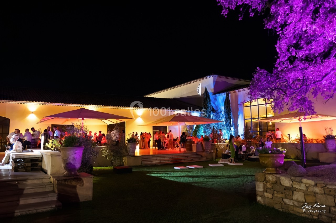 Vue nocturne d'une réception en extérieur avec invités, éclairages colorés et parasols près d'un bâtiment moderne.