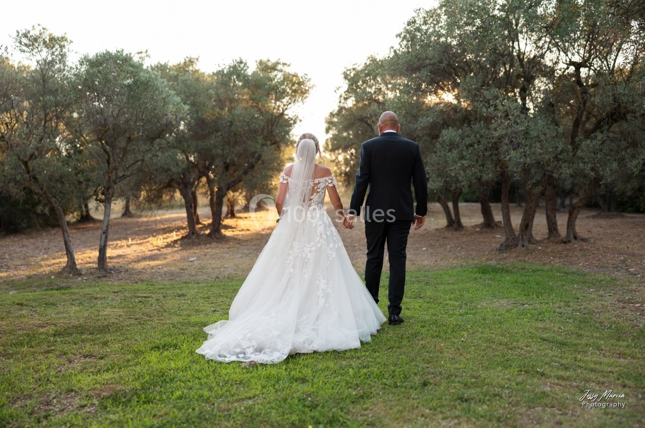 Un couple en tenue de mariage marche main dans la main dans un parc bordé d'arbres au coucher du soleil.