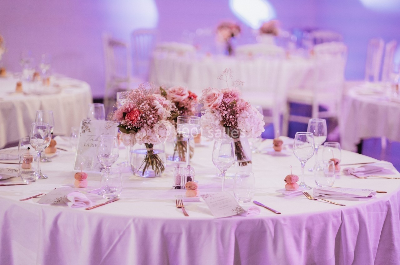 Tables décorées avec des bouquets de fleurs roses et blanches, entourées de chaises, dans une ambiance lumineuse douce.