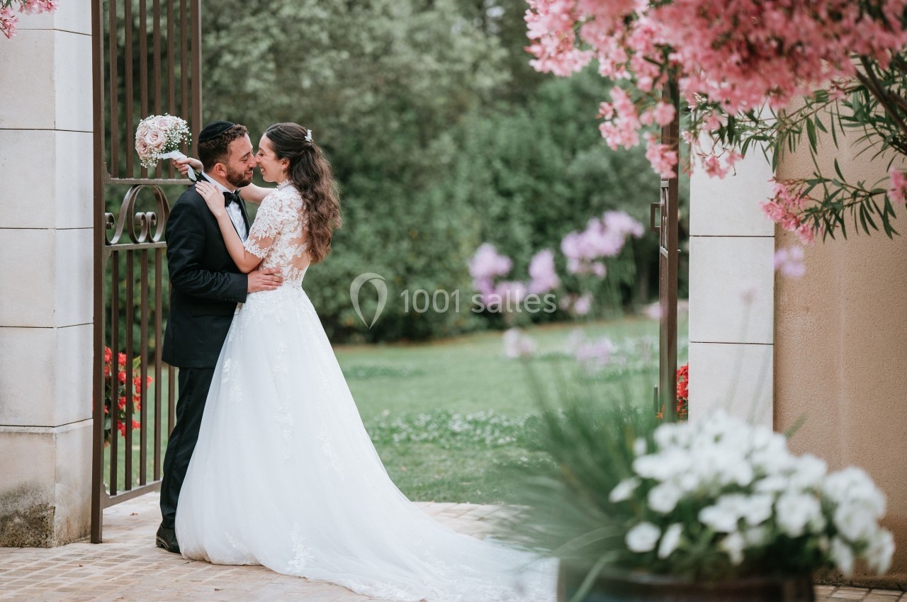 Un couple de mariés s'embrasse devant une porte en fer forgé, entouré de fleurs et d'un jardin verdoyant.