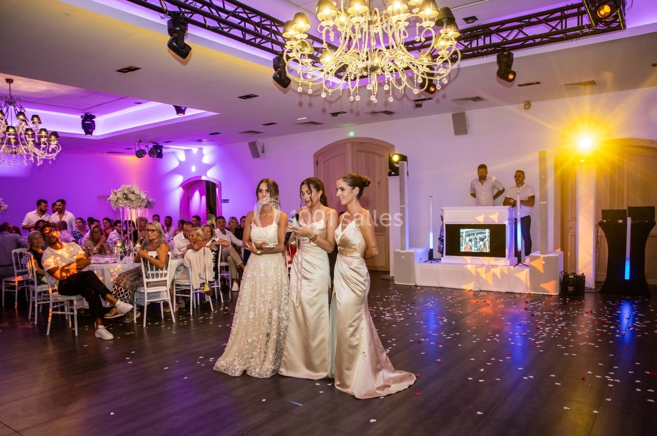 Trois femmes en robes de mariée marchent dans une salle de réception éclairée, entourées d'invités assis.