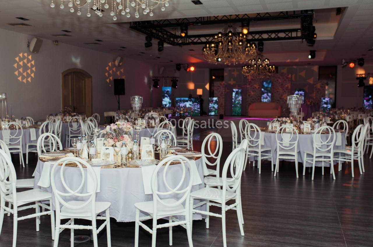 Salle de réception élégante avec tables rondes dressées, chaises blanches et décorations lumineuses suspendues.