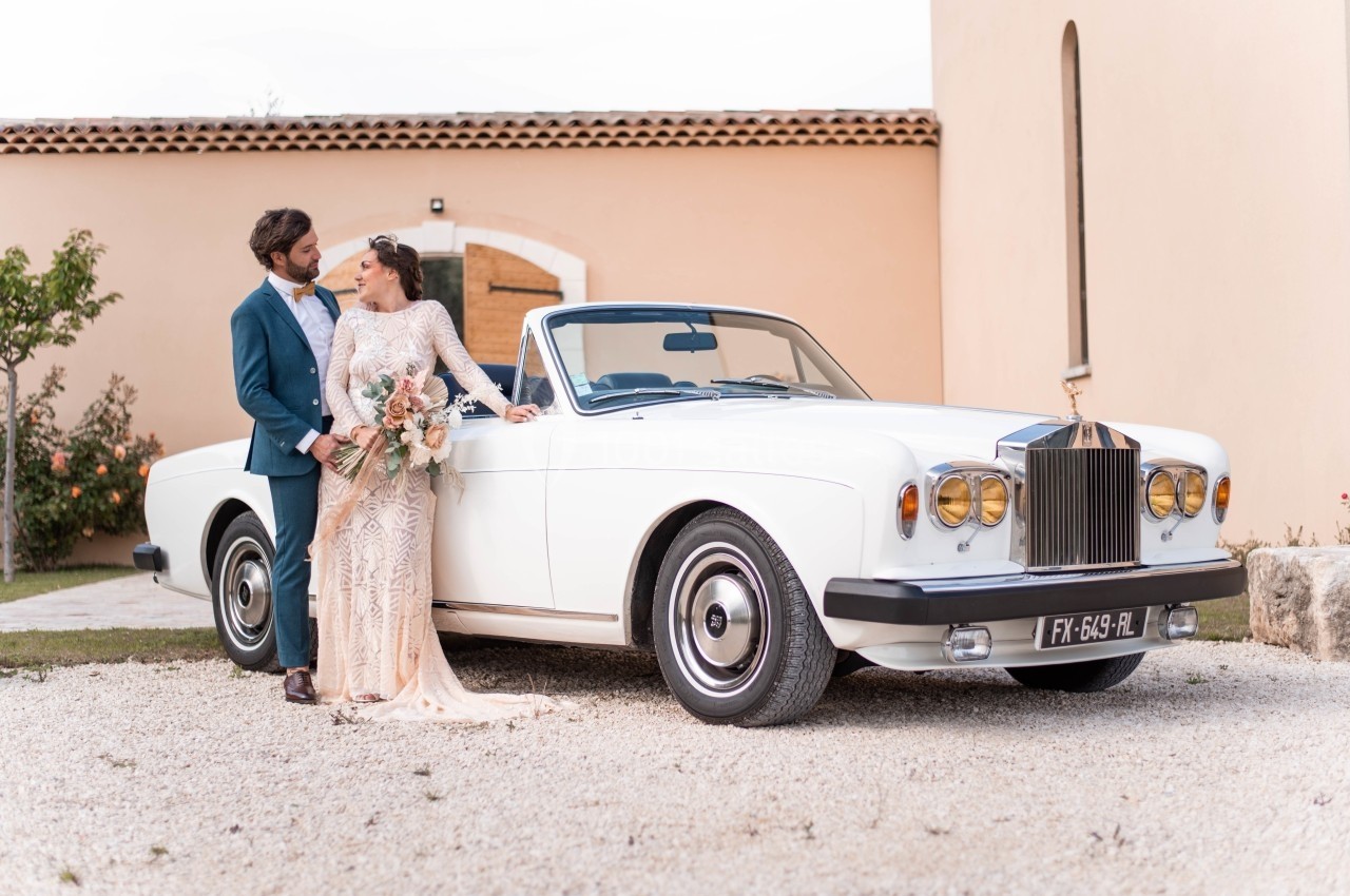 Un couple élégant pose près d'une voiture décapotable blanche devant un bâtiment en pierre.