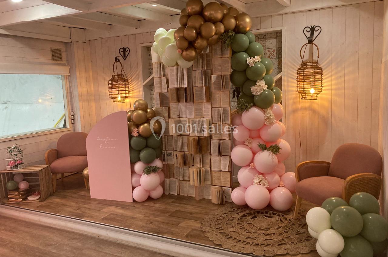 Décor intérieur avec arche de ballons pastel, livres suspendus, fauteuils roses et éclairage chaleureux.