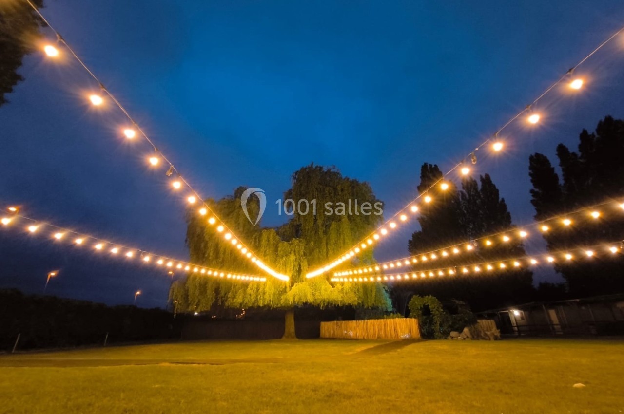 Location salle Hem (Nord) - Le Comte #3 Guirlandes lumineuses suspendues au-dessus d'une pelouse, avec un grand arbre et un ciel crépusculaire en arrière-plan.