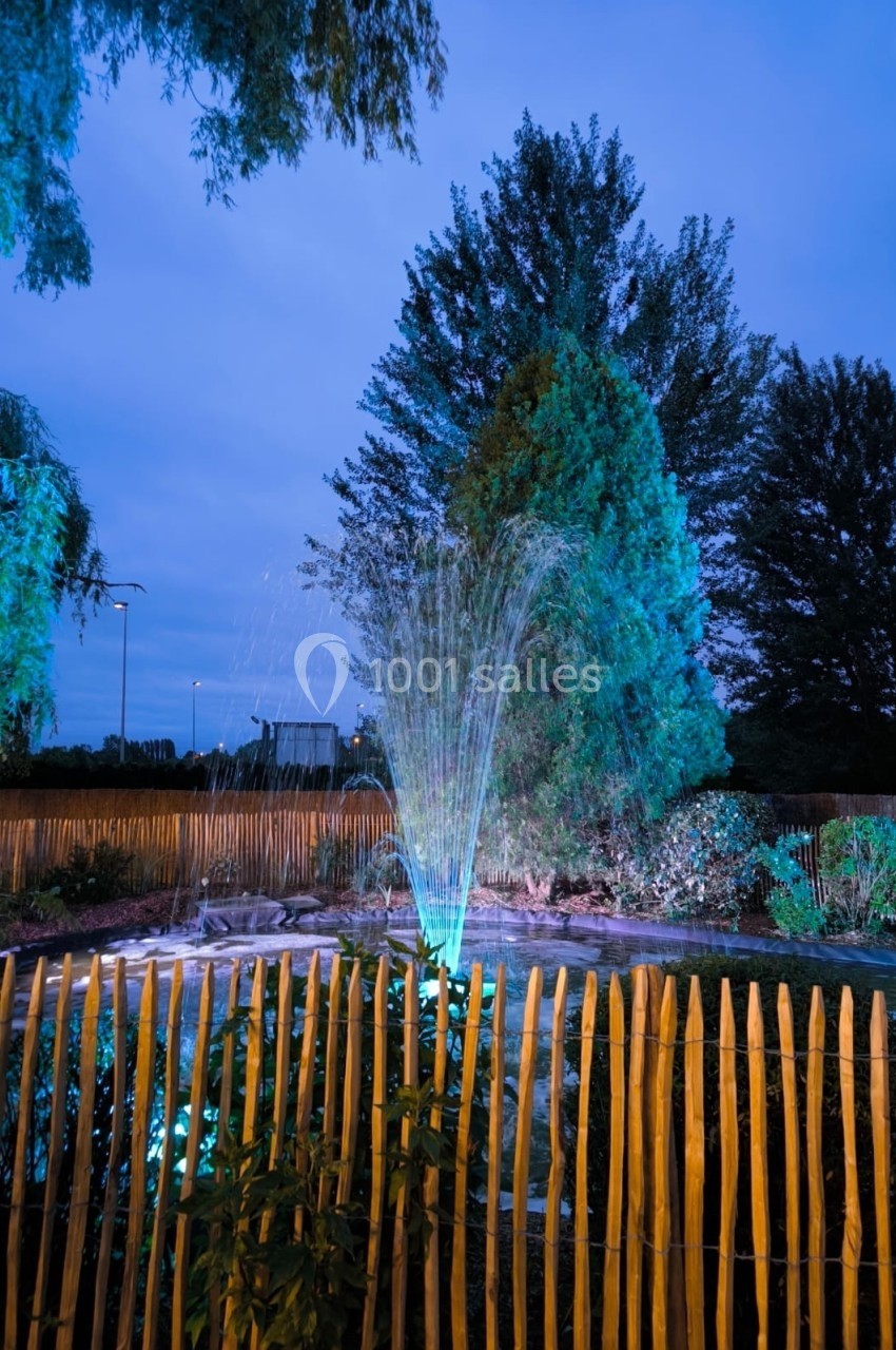 Location salle Hem (Nord) - Le Comte #12 Fontaine éclairée la nuit dans un jardin entouré de palissades en bois, avec des arbres en arrière-plan.