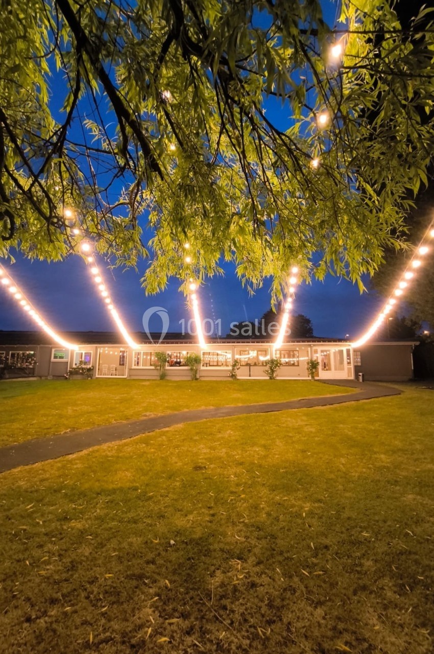 Location salle Hem (Nord) - Le Comte #23 Guirlandes lumineuses suspendues au-dessus d'une pelouse devant un bâtiment éclairé, au crépuscule.