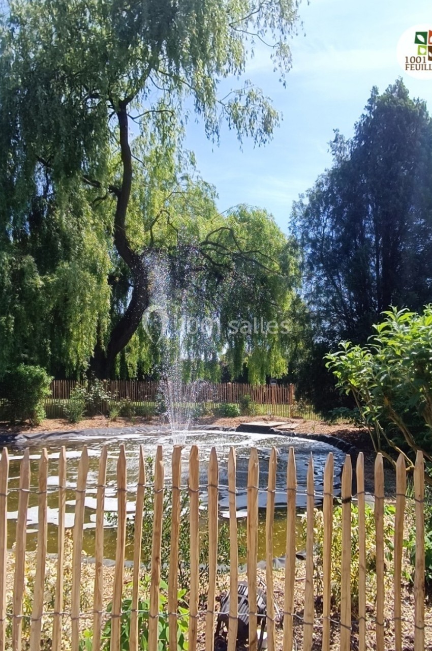 Location salle Hem (Nord) - Le Comte #24 Étang entouré de végétation avec une fontaine centrale, clôturé par une barrière en bois sous un ciel dégagé.