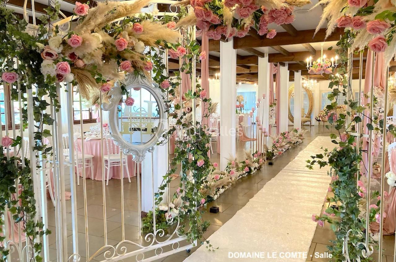 Allée décorée pour un mariage avec arche florale, roses, pampas et chemin blanc dans une salle élégante.