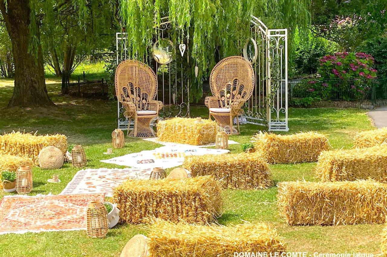 Décoration champêtre avec bottes de paille, tapis, lanternes et fauteuils en osier sous un saule pour une cérémonie en plein…