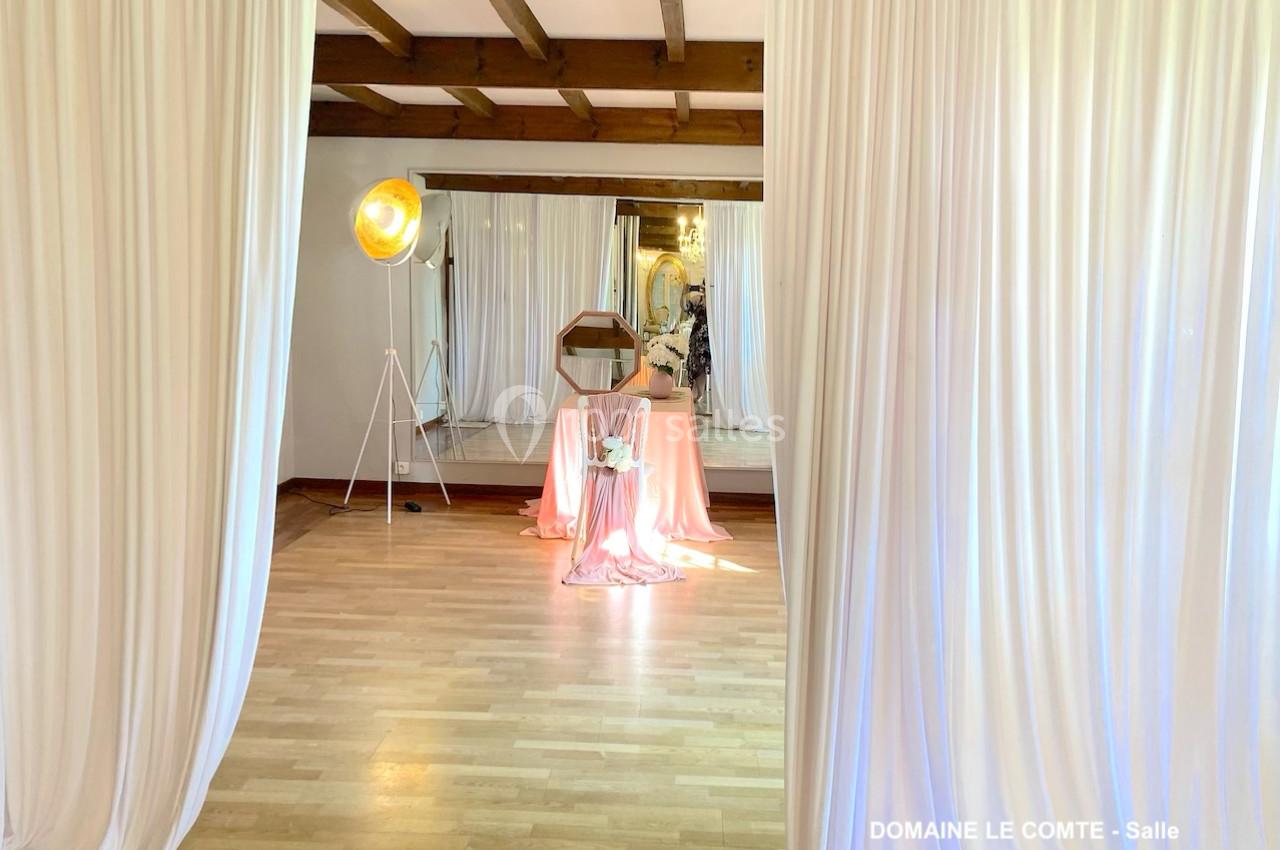 Salle lumineuse avec parquet, rideaux blancs, table décorée de tissu rose et miroir en arrière-plan.