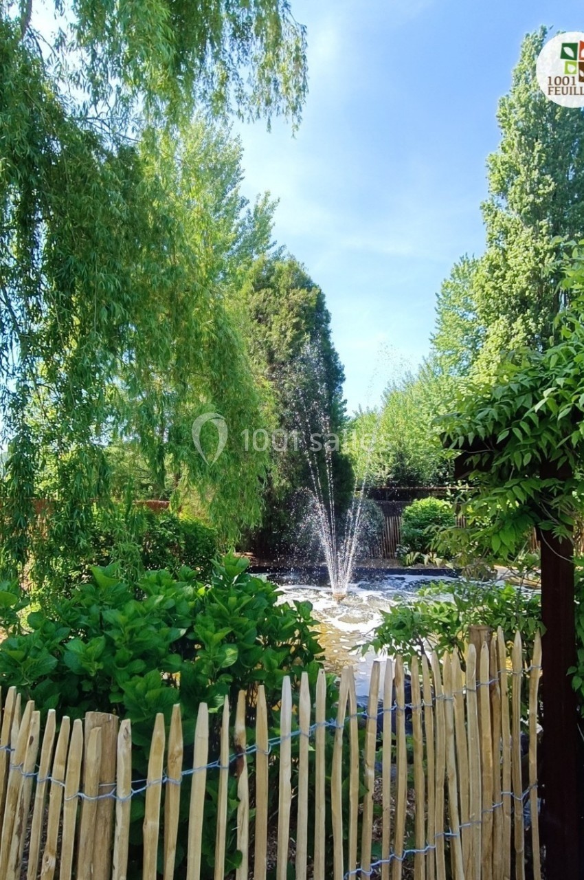 DOMAINE LE COMTE - jusque 200 personnes Un jardin verdoyant avec une fontaine centrale entourée de végétation et une clôture en bois au premier plan.