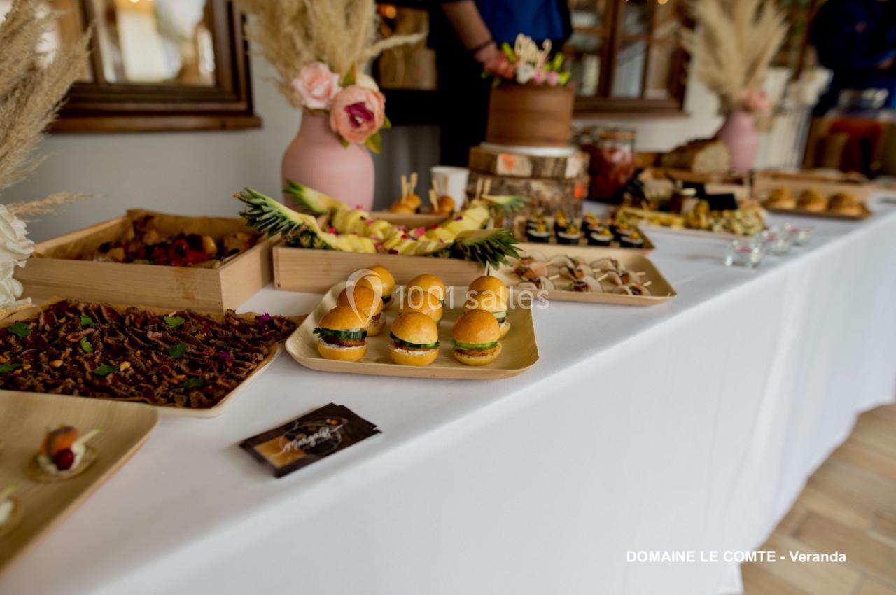 Buffet avec mini-burgers, fruits, desserts et décorations florales sur une table blanche.