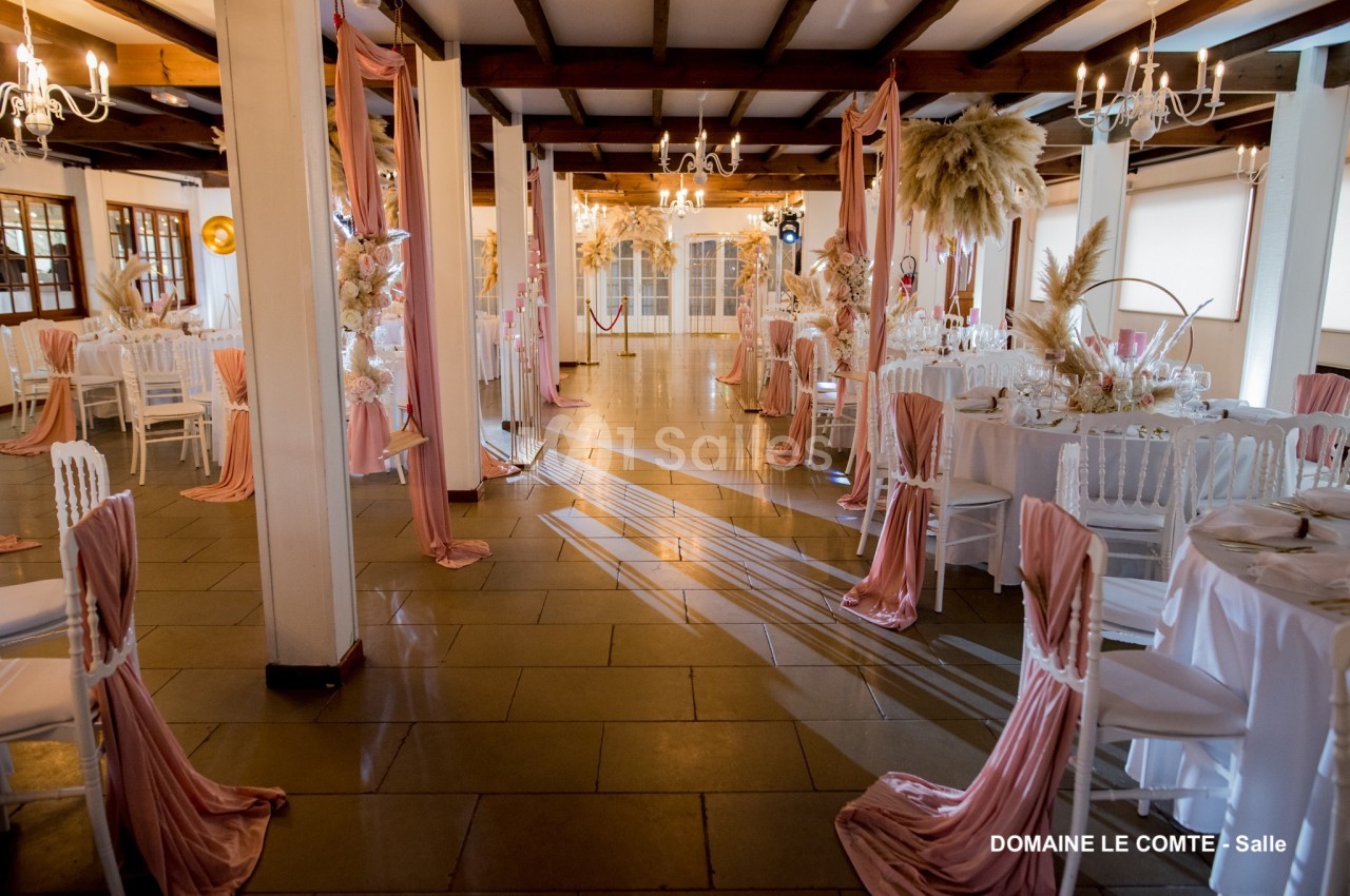 Salle de réception décorée avec des tables rondes, nappes blanches, chaises blanches et drapés roses.