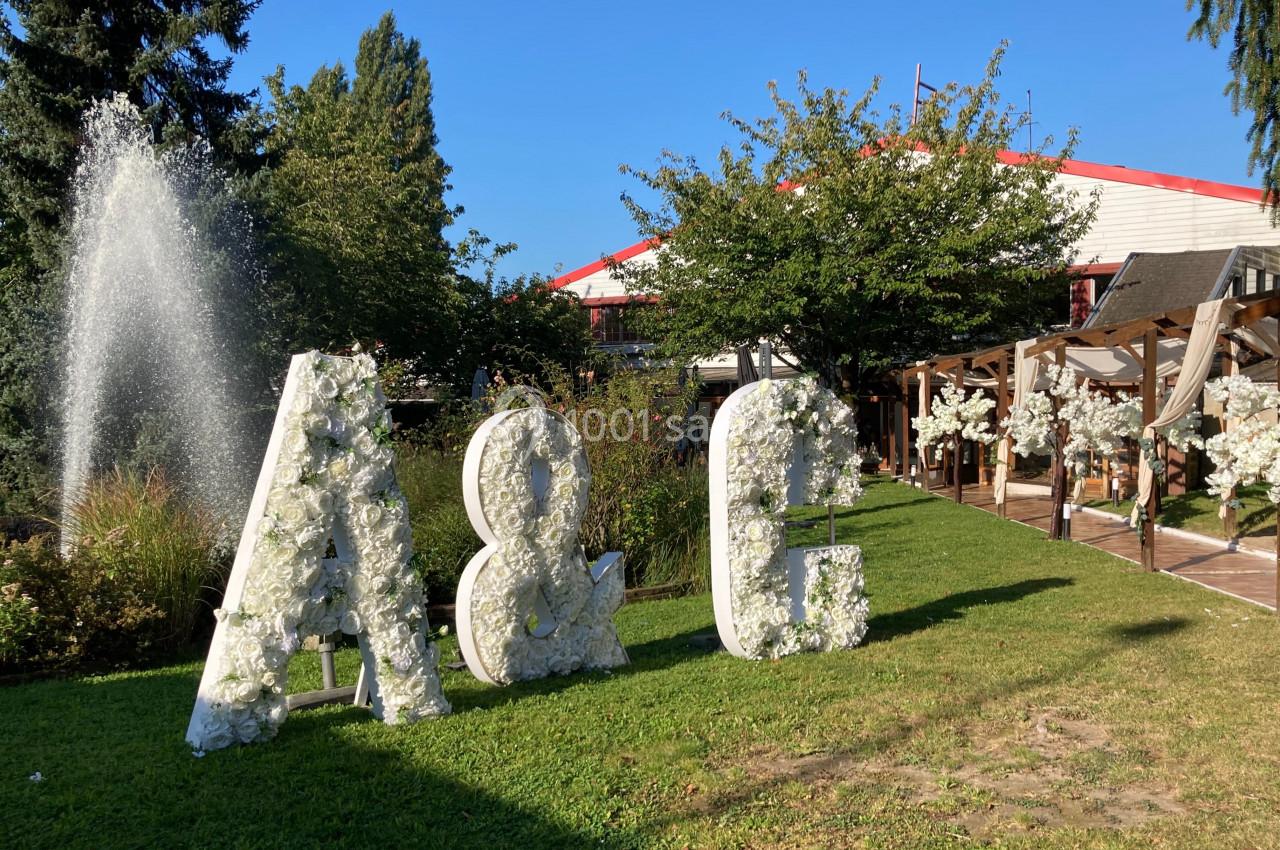 Lettres géantes décorées de fleurs blanches dans un jardin avec fontaine, arbres et bâtiment en arrière-plan.