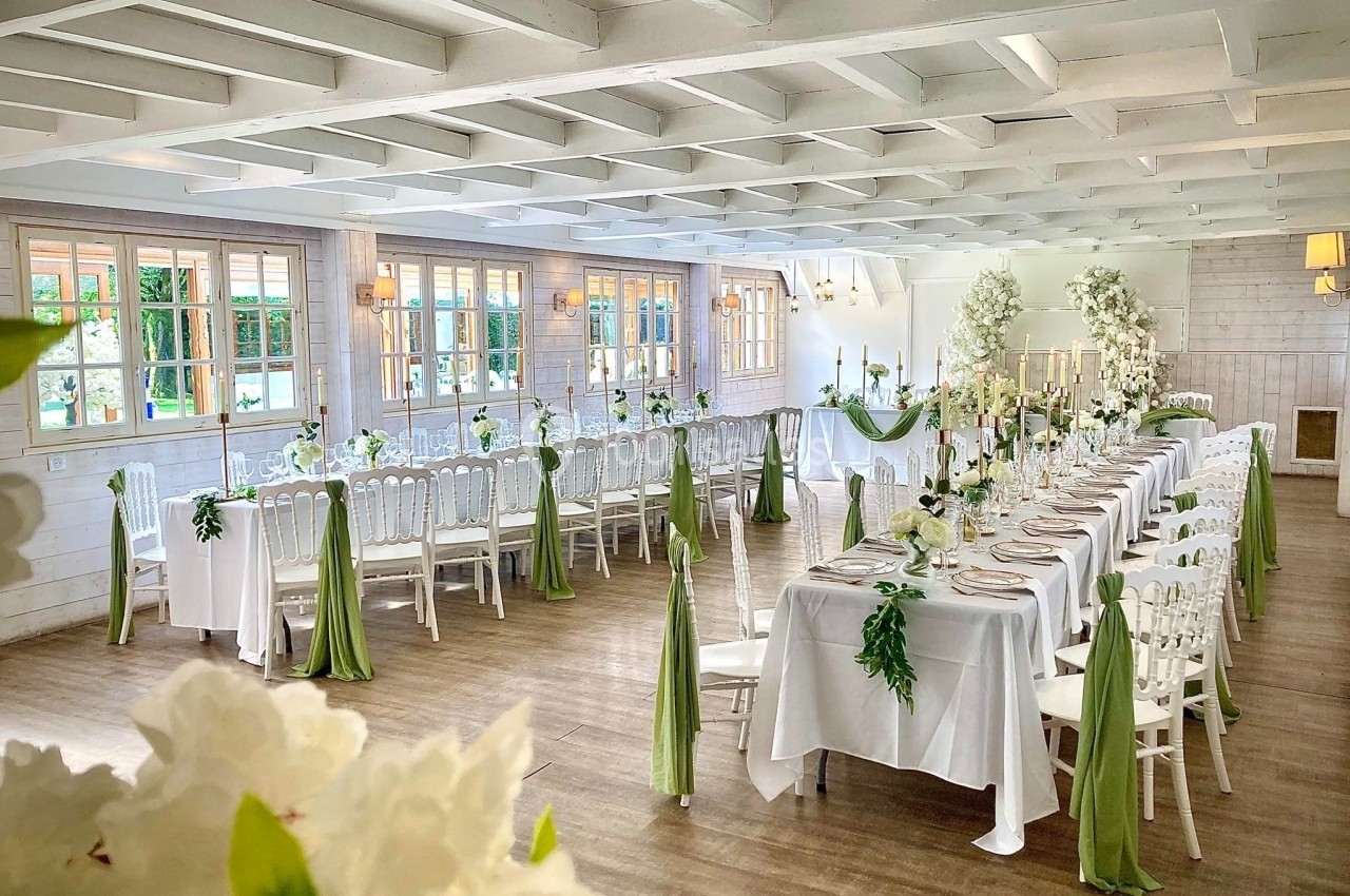 SALLE CHAMPETRE - jusque 100 pers Salle de réception lumineuse décorée pour un mariage, avec longues tables ornées de nappes blanches et touches de vert.