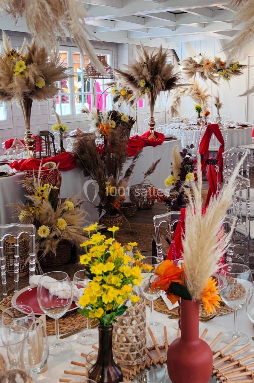 SALLE CHAMPETRE - jusque 100 pers Salle décorée pour un événement avec des tables ornées de fleurs séchées, vases colorés et nappes rouges.