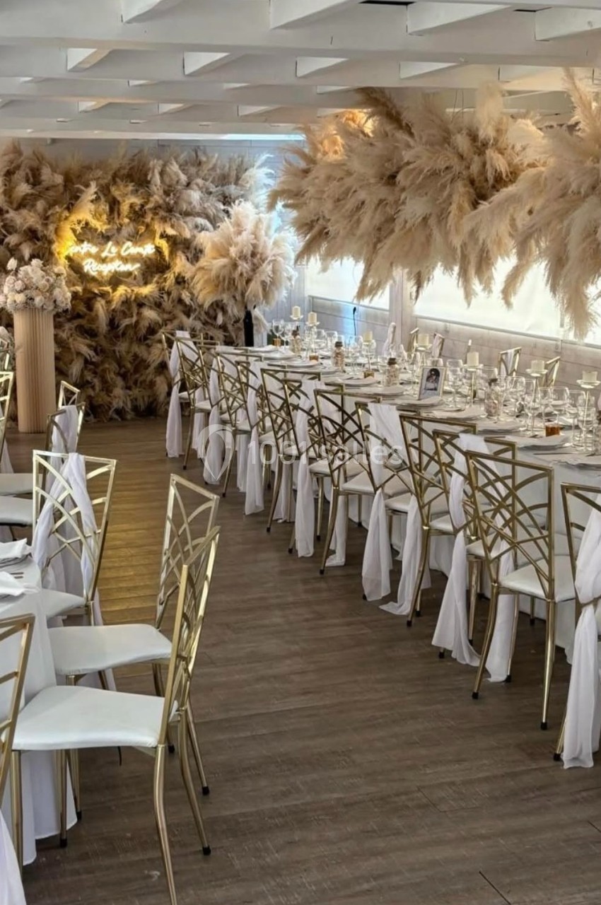 SALLE CHAMPETRE - jusque 100 pers Salle de réception décorée avec des pampas, tables dressées avec nappes blanches et chaises ornées de rubans.