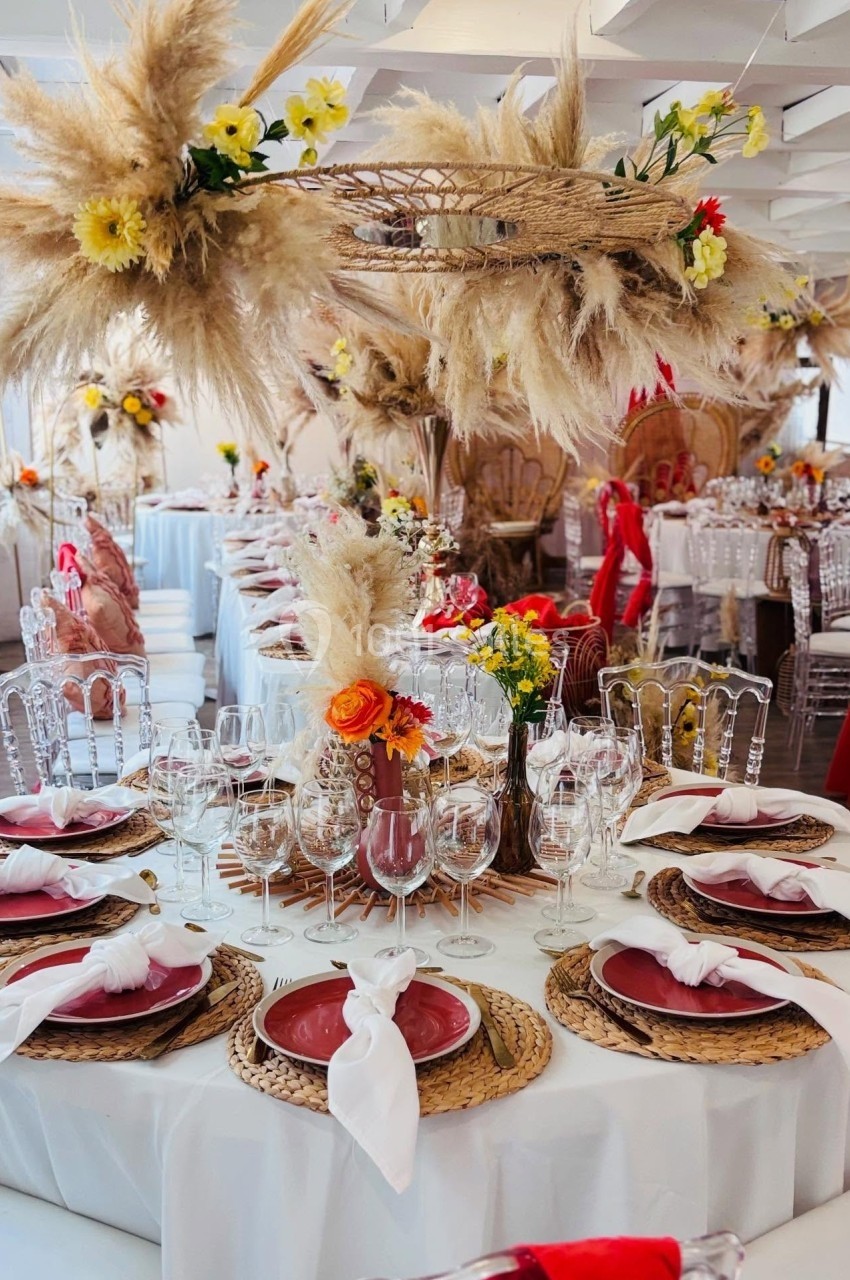 SALLE CHAMPETRE - jusque 100 pers Table décorée pour un événement avec des assiettes rouges, des serviettes blanches et des arrangements de fleurs et pampas.