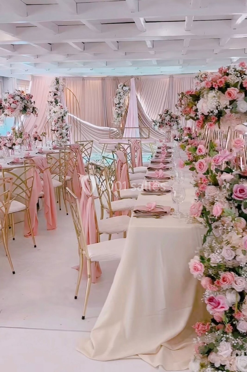 SALLE CHAMPETRE - jusque 100 pers Salle de réception décorée avec des tables élégantes, nappes blanches et arrangements floraux roses et blancs.