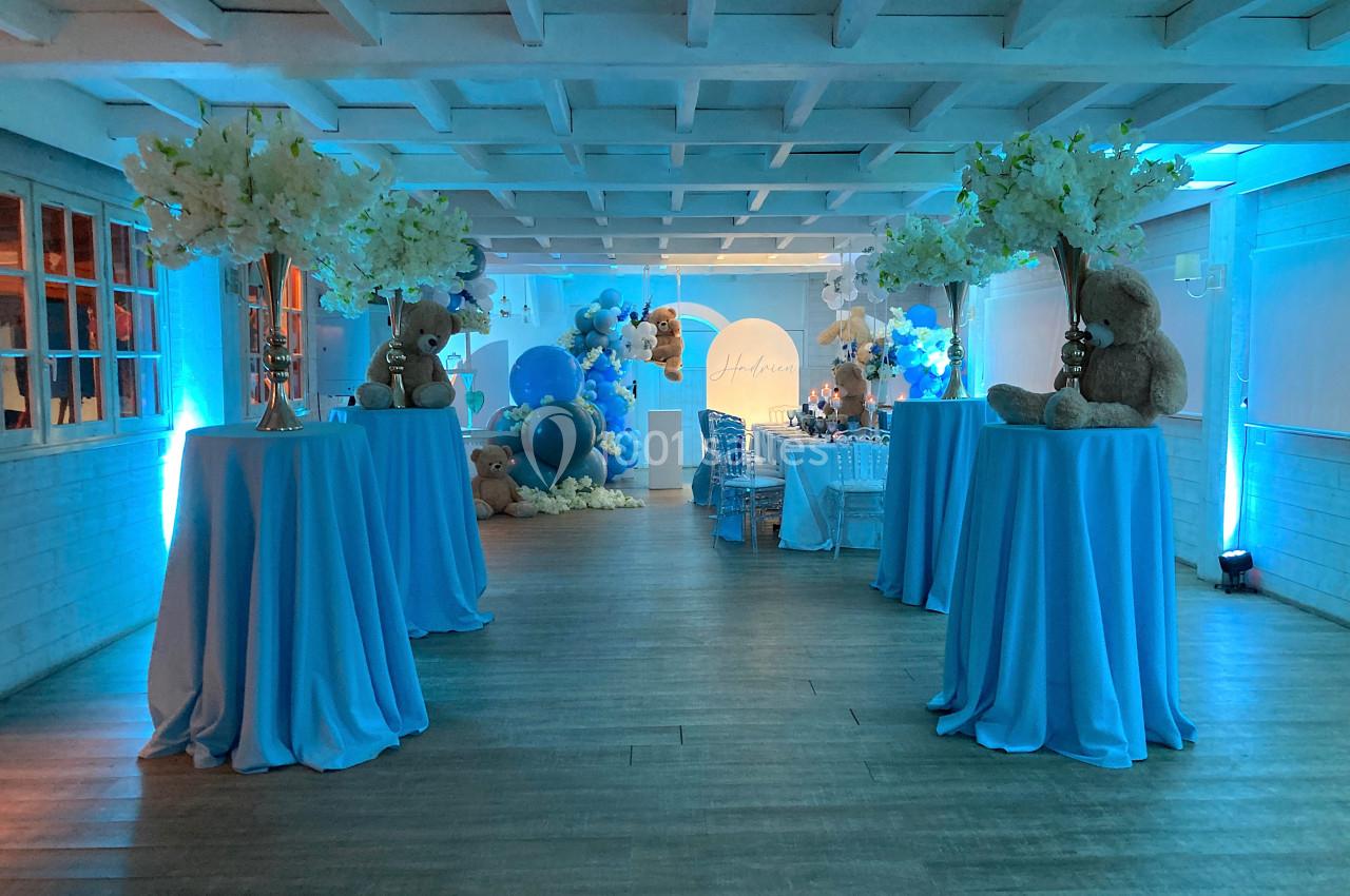 Salle décorée en bleu avec tables hautes, fleurs blanches, ballons et peluches dans une ambiance festive.