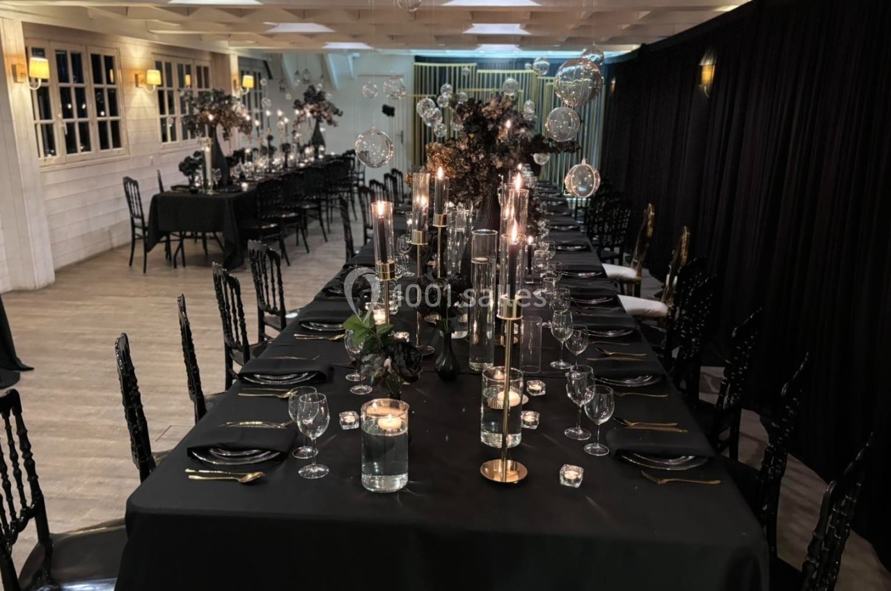 SALLE CHAMPETRE - jusque 100 pers Salle de réception élégante avec longues tables noires décorées de bougies, verrerie et arrangements floraux sombres.
