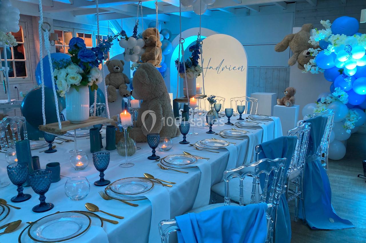 Table décorée pour une fête avec vaisselle élégante, ours en peluche, ballons bleus et bougies dans une ambiance lumineuse.