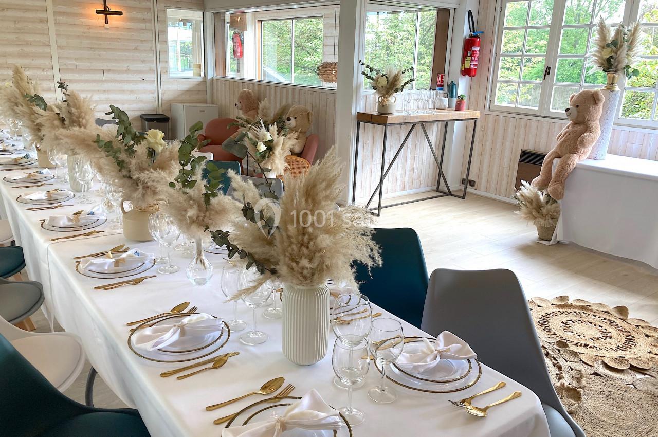 SALLE COSY - 80 PERS - Location à la journée 8h-18h Salle lumineuse décorée pour un événement, avec une longue table dressée, des fleurs séchées et des ours en peluche.