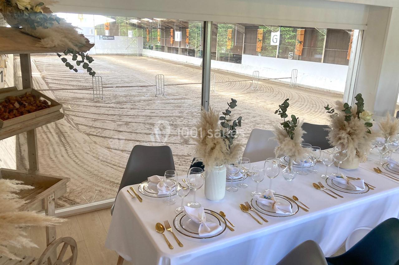 SALLE COSY - 80 PERS - Location à la journée 8h-18h Table élégamment dressée avec vaisselle blanche et décorations florales, donnant sur une piste équestre en arrière-plan.