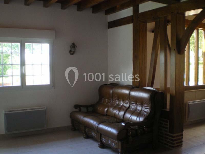 Canapé en cuir marron dans un salon avec poutres en bois, mur blanc et fenêtre laissant entrer la lumière naturelle.