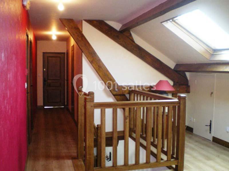 Escalier en bois avec rambarde, sous des poutres apparentes, éclairé par une fenêtre de toit et des lumières.