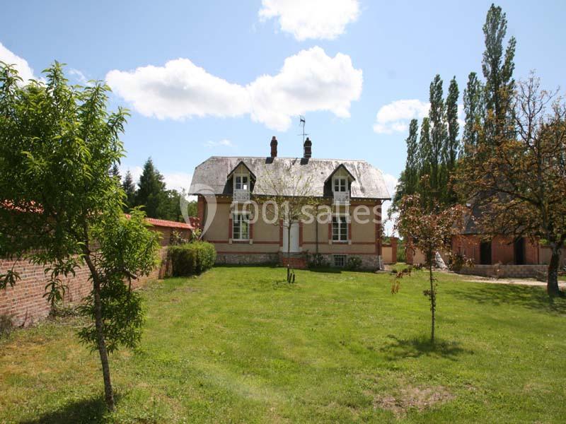 Maison en briques avec toit en ardoise, entourée d'un jardin verdoyant et d'arbres sous un ciel partiellement nuageux.