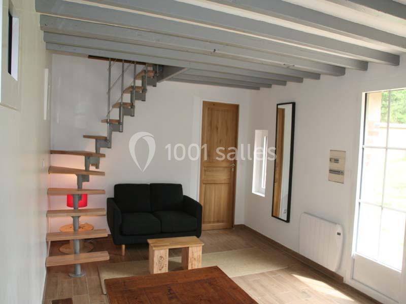 Salon avec escalier en bois et métal, canapé noir, table basse en bois, mur blanc et plafond avec poutres apparentes.