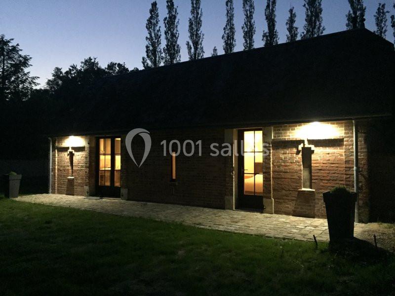 Façade d'une maison en briques éclairée par des appliques murales, avec des portes vitrées et un jardin au premier plan.