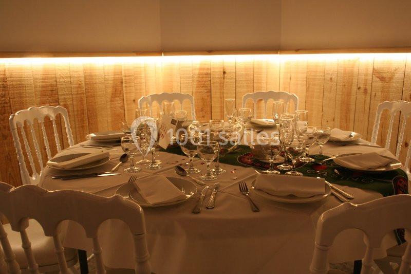 Table ronde dressée avec nappes blanches, chaises assorties, vaisselle et verres, éclairée par une lumière tamisée.