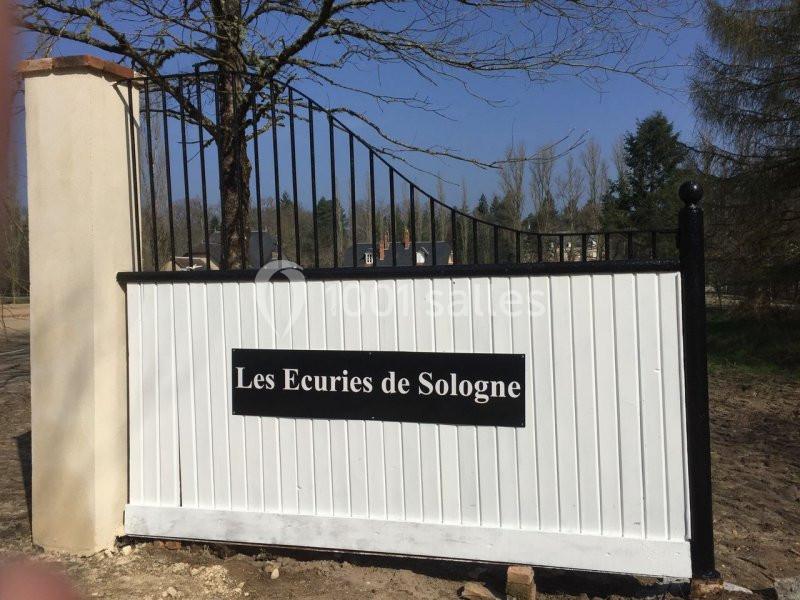 Portail blanc et noir avec un panneau indiquant ’Les Écuries de Sologne’, entouré d'arbres et d'un ciel dégagé.