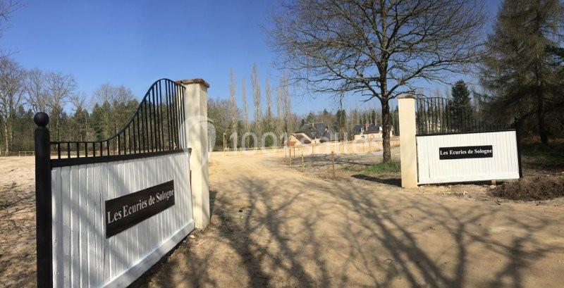 Entrée d'une propriété avec des portails blancs et noirs, entourée d'arbres et d'un chemin en terre.