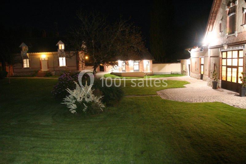 Cour extérieure éclairée la nuit, avec pelouse, arbustes et bâtiments en briques aux fenêtres illuminées.